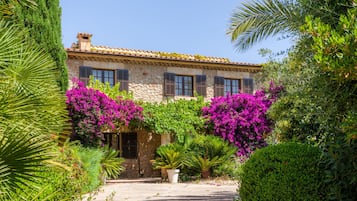 A stone building with a tiled roof, a central entrance, and lush greenery including purple bougainvillea.