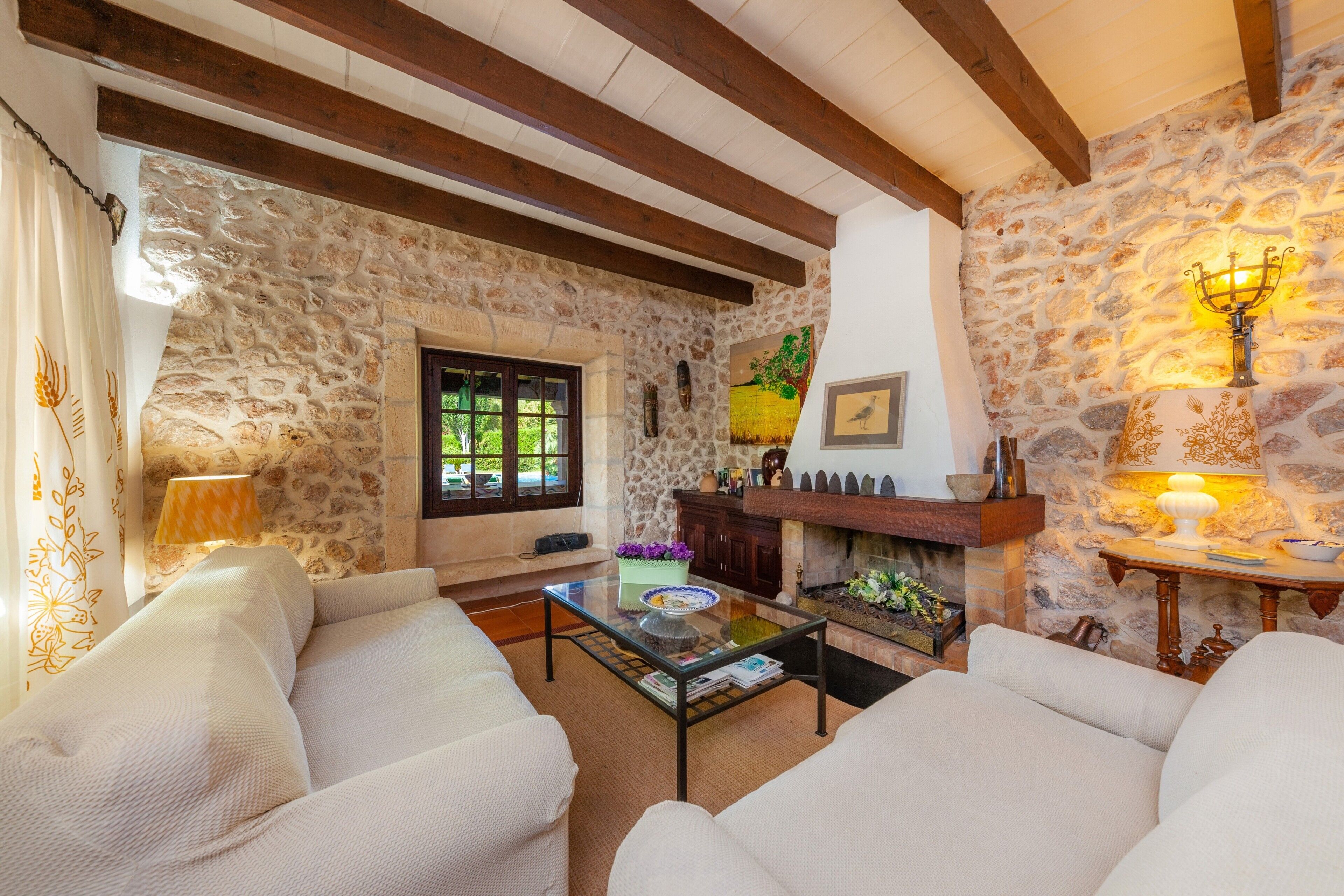 A cozy living room with stone walls, wooden beams, a fireplace, and a glass coffee table.