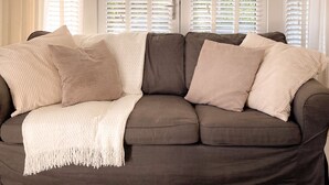 A living room with a dark brown sofa, white curtains, and a window with blinds. - Secluded Countryside Lodge on 25 Acres | Private Deck & Views | Kent Escape (Biddenden)