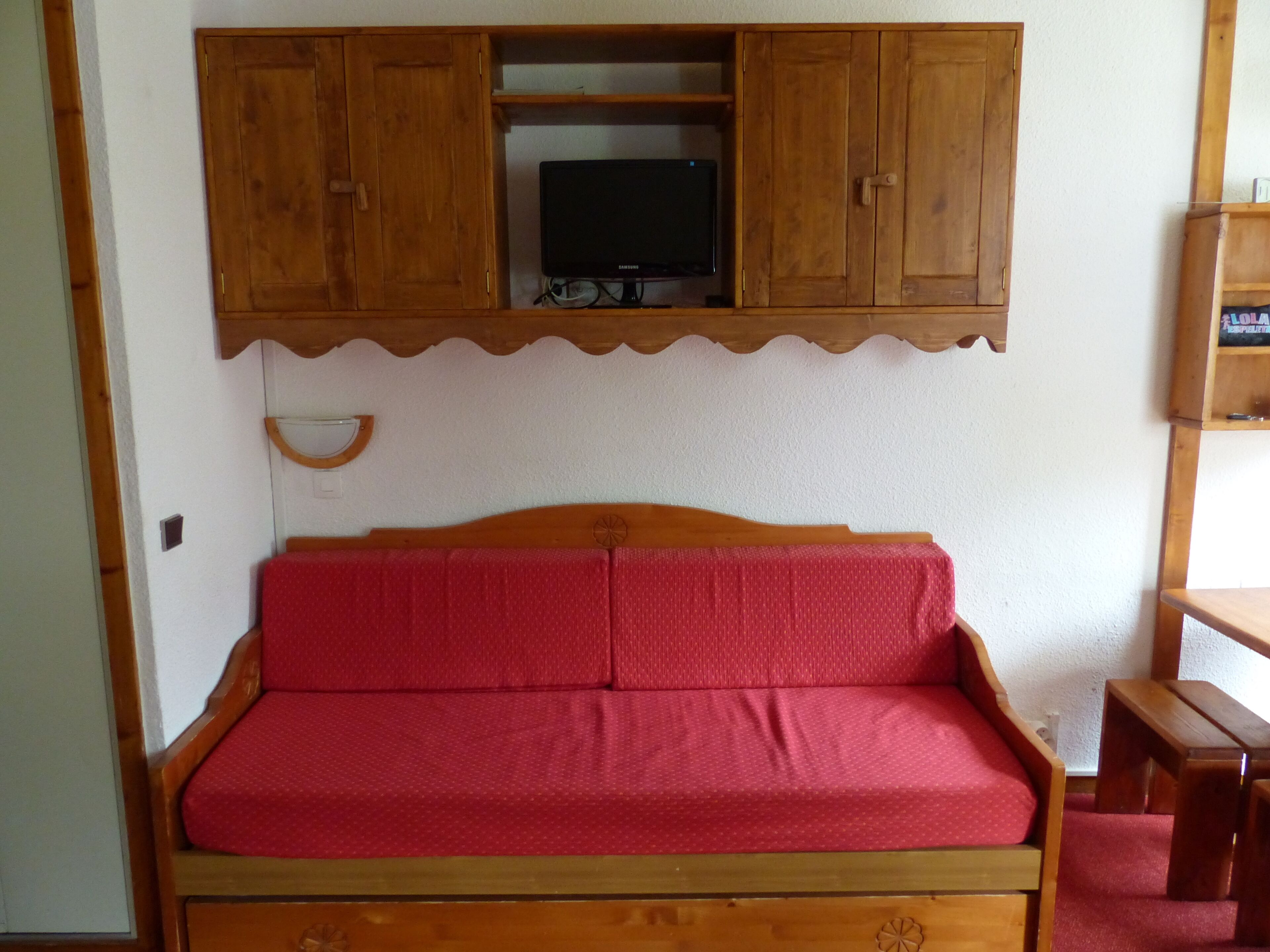 A wooden bed with a red mattress, a wooden cabinet above it, and a television on a shelf.