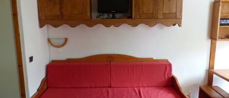 A wooden bed with a red mattress, a wooden cabinet above it, and a television on a shelf.