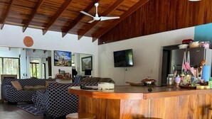 A spacious living area with a wooden ceiling, a sofa, a TV, and a kitchen area with stools.