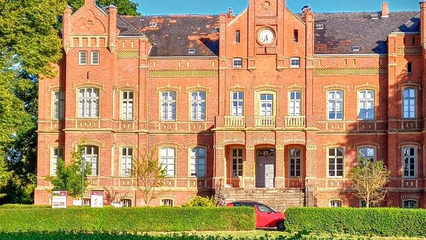 A large red brick building with a clock tower, multiple windows, and a red car parked in front.
