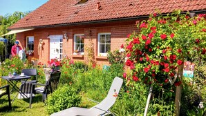 A brick house with a red-tiled roof, a garden with red flowers, and a table with chairs.