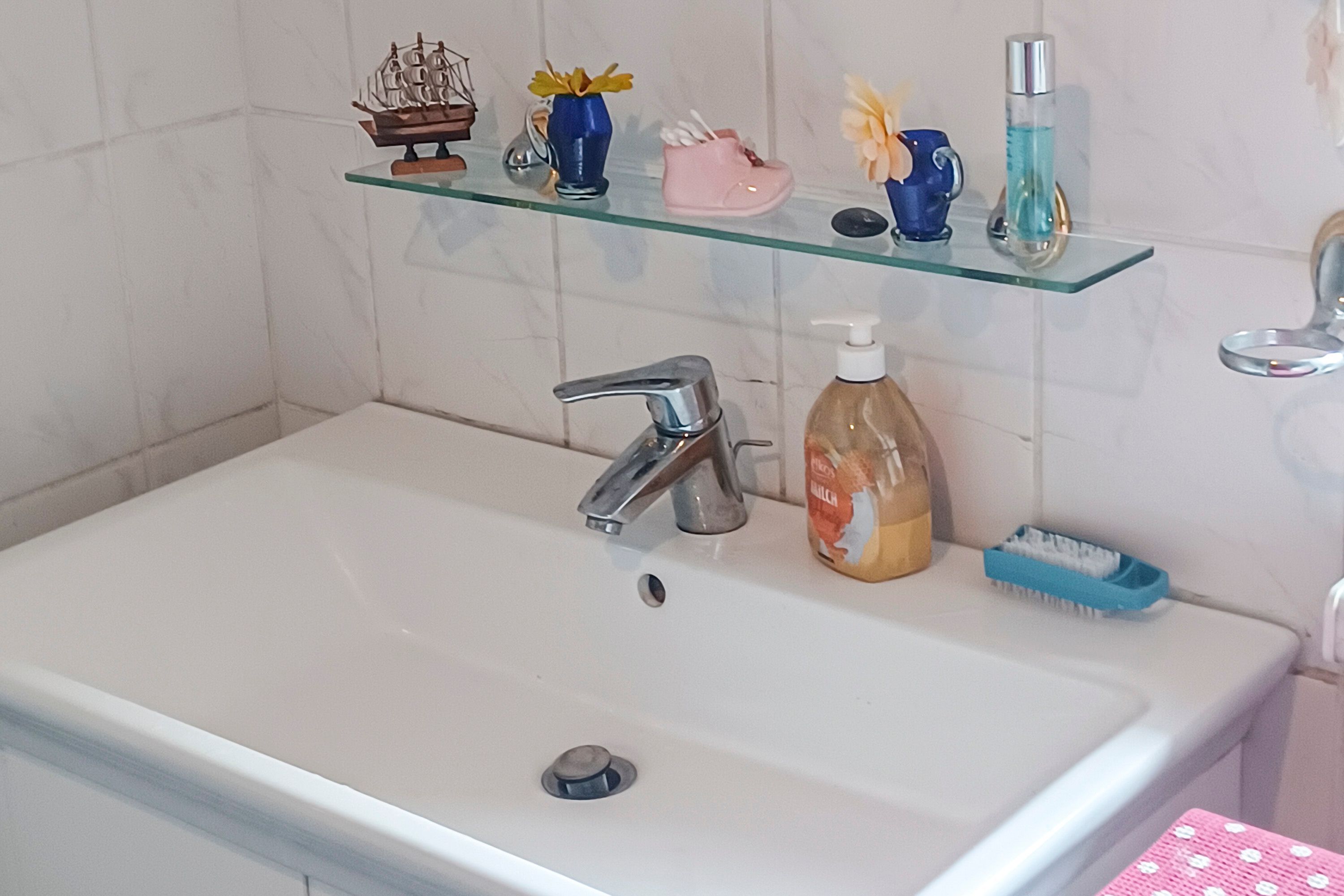 Un baño con un lavamanos blanco, un modelo de barco, un florero con flores amarillas, un zapato rosa, una botella azul y un dispensador de jabón.