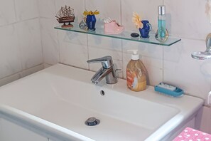 Een badkamer met een witte wasbak, een scheepsmodel, een vaas met gele bloemen, een roze schoen, een blauwe fles en een zeepdispenser.