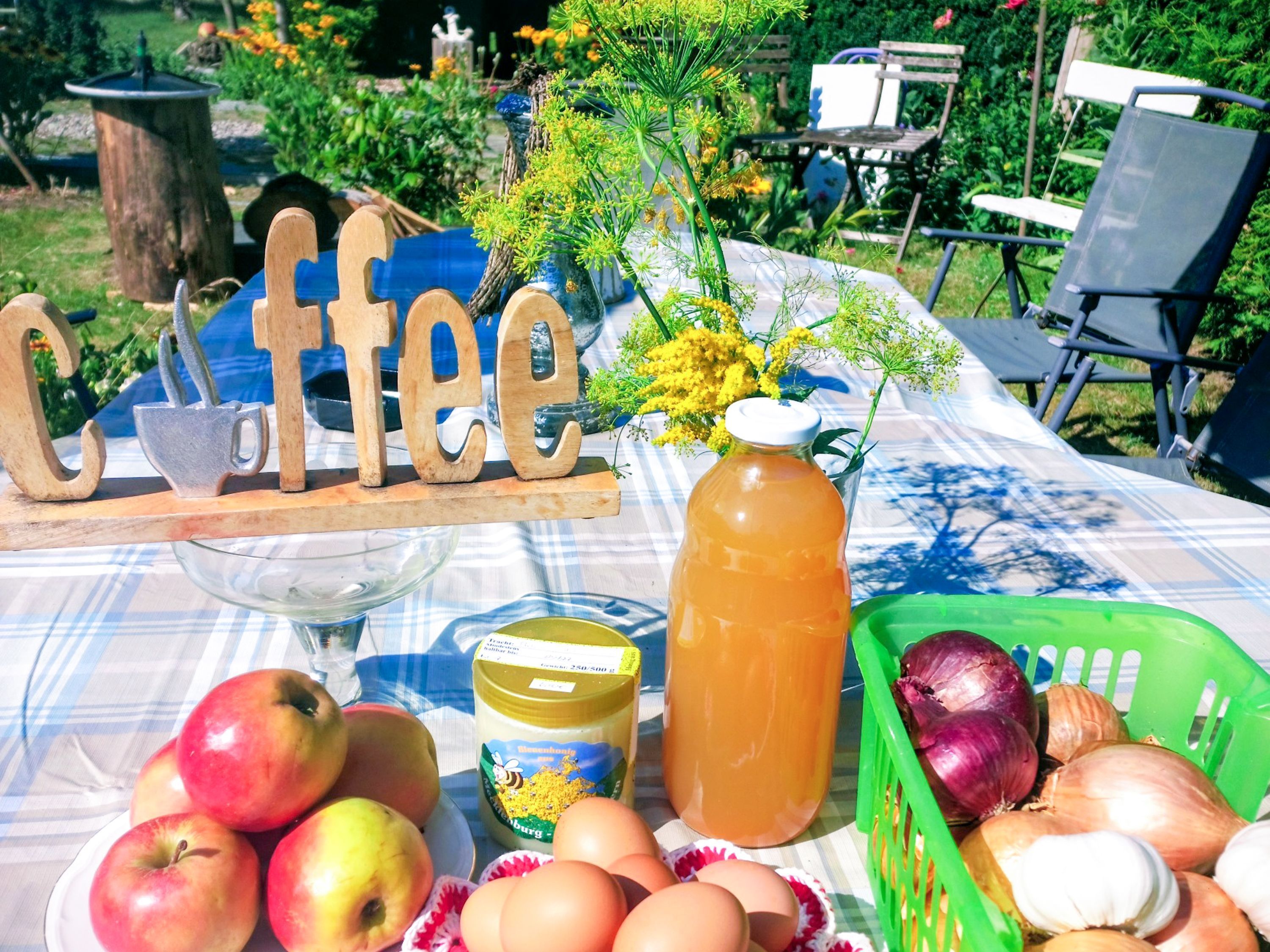 A table with a coffee sign, apples, eggs, a honey jar, and a green basket with onions.
