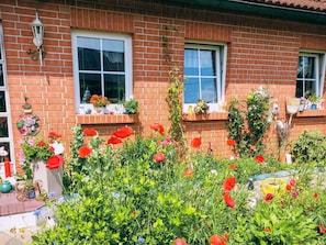 Een bakstenen huis met twee ramen, een klein portaaltje en een tuin met rode bloemen.