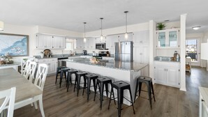 A modern kitchen with white cabinets, a large island with bar stools, stainless steel appliances, and pendant lighting. - "Good Times and Tan Lines " - Luxury Oceanfront Home w/ Private Pool, 6 Bedrooms / 5 Bathrooms (Holden Beach)