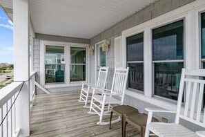 A covered porch with white rocking chairs and a small table, large windows with shutters, and a view of the street. - "Holden Mystique" - 2nd Row Home w/ Pool, 4 Bedrooms / 3 Bathrooms (Holden Beach)