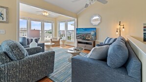 Ein geräumiges Wohnzimmer mit einer großen Couch, einem Couchtisch und einem Fernseher. Es gibt einen Deckenventilator, ein Fenster mit Ausblick und eine Wandlampe.