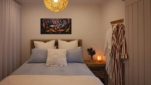 A bedroom with a bed, pillows, a nightstand with a salt lamp, a painting, and a robe hanging on the wall. - Modern Unit/Beach Walk/Drive to Winery (Port Noarlunga South)