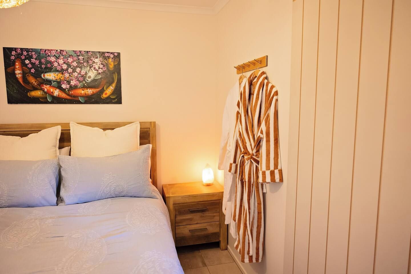 A bedroom with a bed, pillows, a nightstand with a lamp, a robe hanging on a rack, and a painting on the wall.