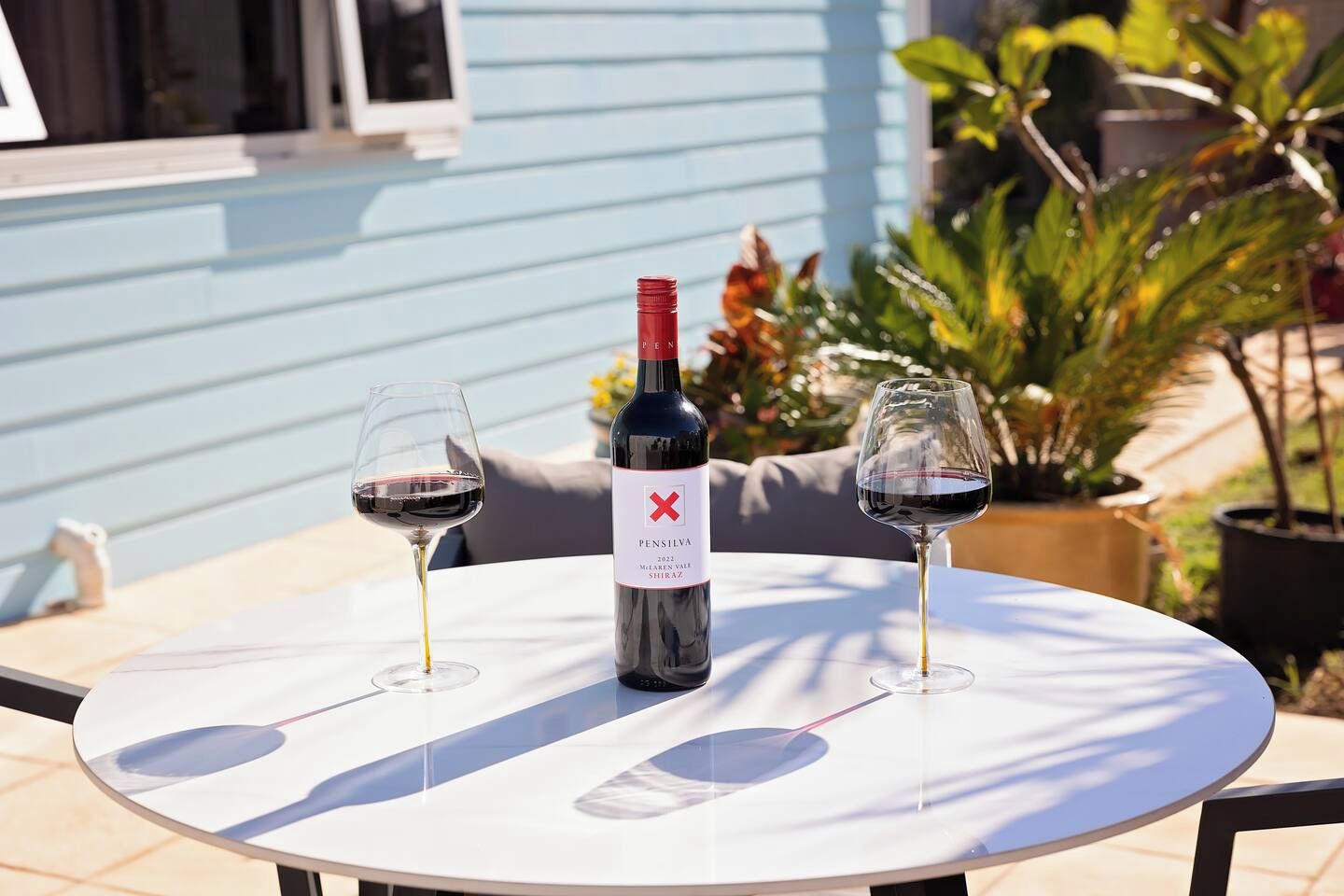 Una mesa con una botella de vino y dos copas, colocada al aire libre frente a un edificio azul y plantas en el fondo.