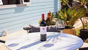 Una mesa con una botella de vino y dos copas, colocada al aire libre frente a un edificio azul y plantas en el fondo.