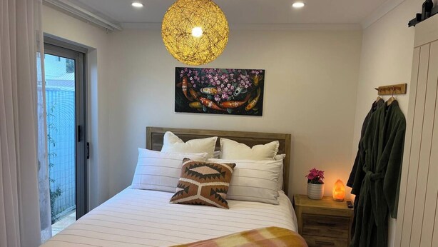 A bedroom with a bed, a hanging light fixture, a painting, a nightstand, a robe, and a sliding door. - Modern Unit/Beach Walk/Drive to Winery (Port Noarlunga South)