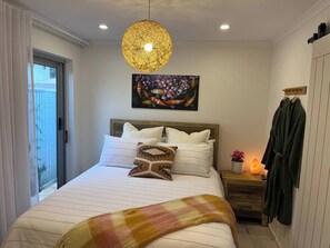 A bedroom with a bed, a hanging light fixture, a painting, a nightstand, a robe, and a sliding door.