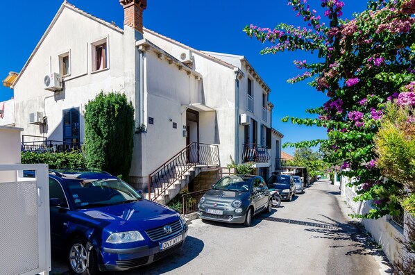 Une rue étroite bordée de voitures garées et un immeuble blanc doté d’un balcon.