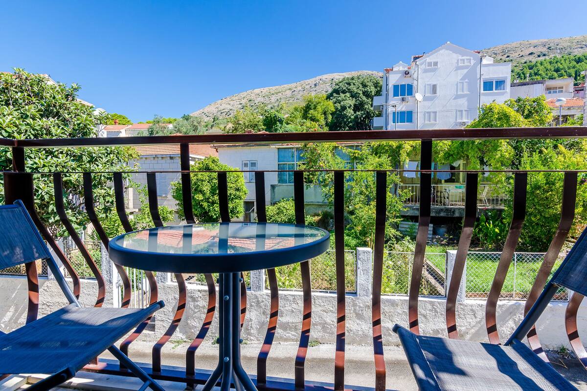 A balcony with a glass table and two chairs, overlooking a hillside with buildings.