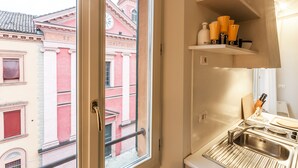 Una cocina con vista a la calle, donde se ve un edificio con columnas y un reloj. Hay estantes con utensilios de cocina y un lavamanos con grifo.