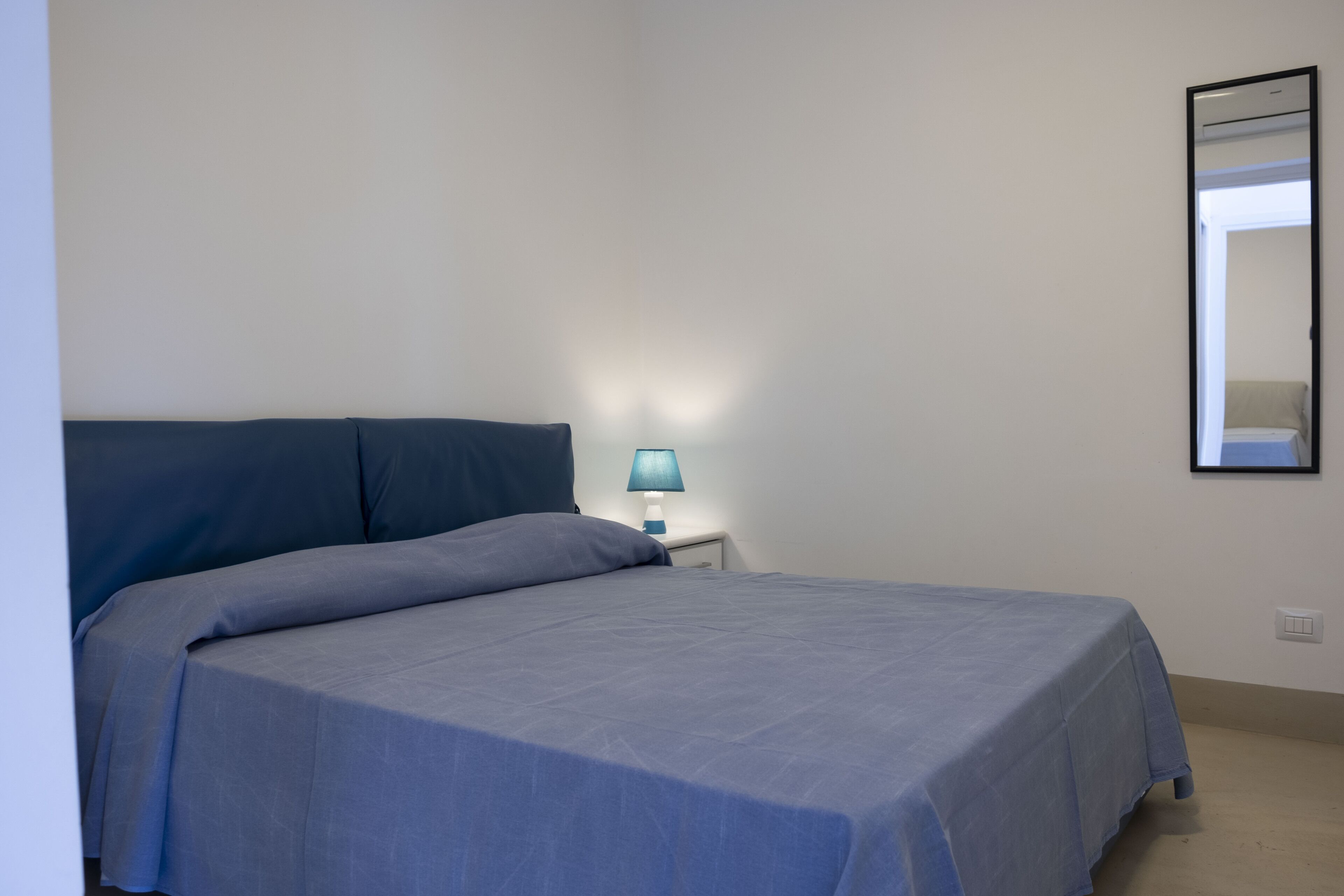 Een net opgemaakt bed met een blauw beddegoed en kussens, een bedlampje en een spiegel aan de muur.