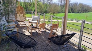 An outdoor patio with wicker furniture, a table with a bottle and glasses, and a view of a green field and trees.