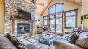Una sala de estar acogedora con chimenea de piedra, sofás de cuero, un televisor de pantalla plana y amplios ventanales con vistas al exterior.
