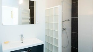 Un baño moderno con lavamanos blanco, espejo, toallero y zona de ducha con azulejos oscuros.