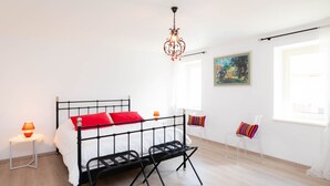 Un dormitorio moderno con estructura de cama metálica negra, ropa de cama blanca y almohadas rojas. Hay una mesita de noche con una lámpara, una silla de plástico transparente y un cuadro en la pared.
