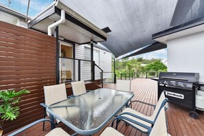 An outdoor patio with a glass table and chairs, a barbecue grill, and a wooden deck.