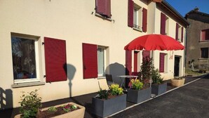 Un bâtiment avec des volets rouges, des tables et chaises à l’extérieur sous un parasol rouge, et des jardinières contenant différentes plantes.