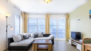 A living room with a sectional sofa, a coffee table, a TV on a stand, and a floor lamp. - (2) Barbara, 3-room apartment, wheelchair accessible, ground floor - Ostseeferienhaus (Am Salzhaff)
