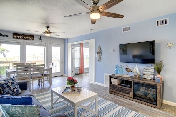 A living room with a TV, ceiling fans, a wooden entertainment center, and a view through a door. - Gulf Getaway! Ocean View, Pool, Boardwalk, Beach (Port Aransas)