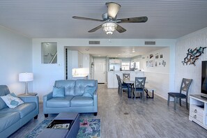 Un salon spacieux avec un canapé bleu, une table basse et un ventilateur de plafond. On y trouve également un coin repas avec des chaises et une table, ainsi qu’une cuisine à l’arrière-plan.