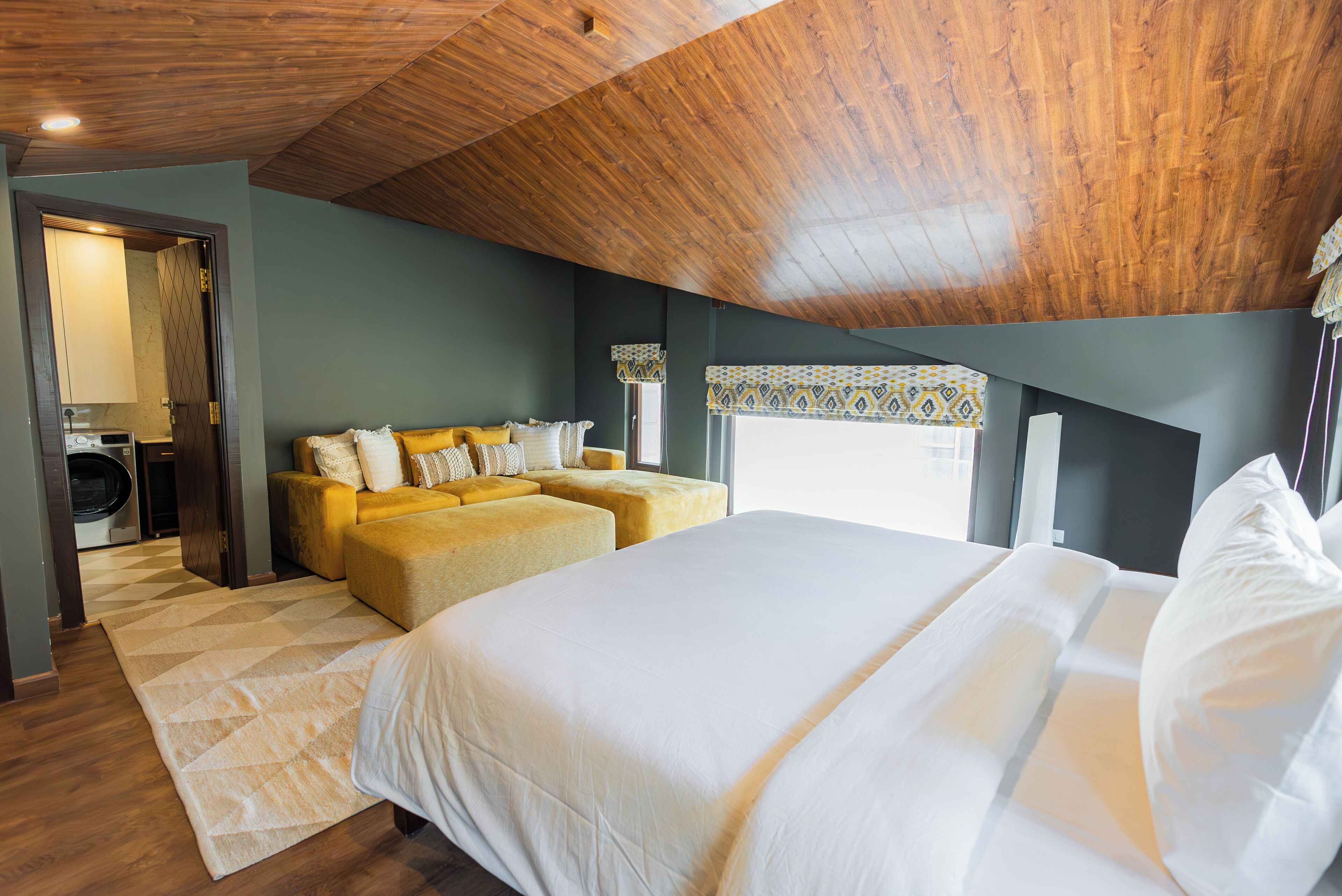Un dormitorio con una cama grande, un sofá y una mesita. Se ve una ventana con cortinas estampadas y un techo de madera.
