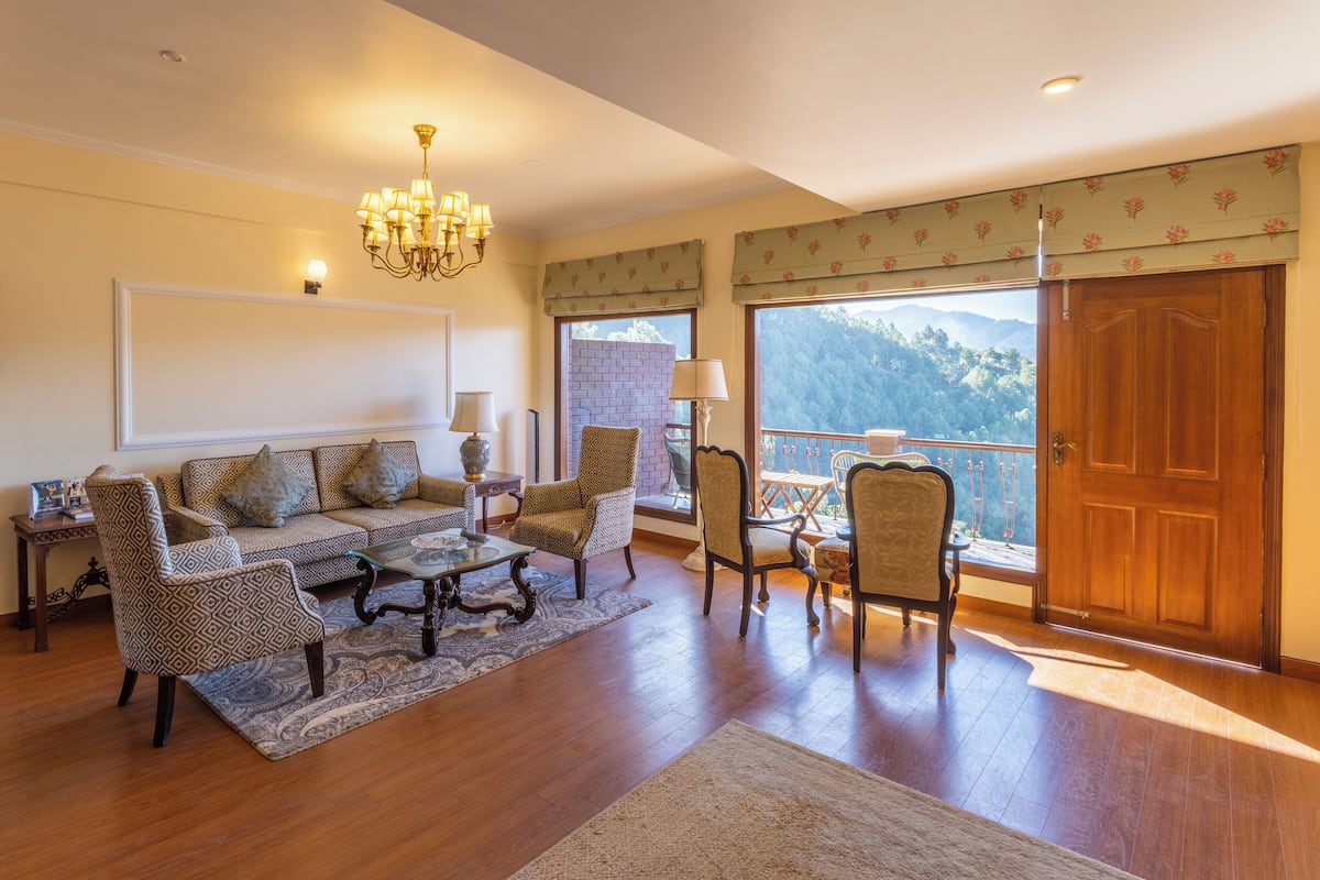A spacious living room with a sofa set, patterned chairs, a chandelier, and a view of a mountainous landscape through large windows.