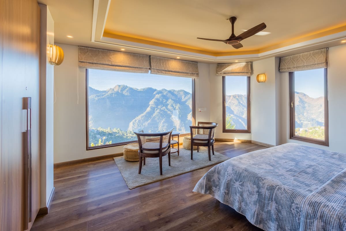 A bedroom with a large window, a bed, two chairs, a ceiling fan, and a view of mountains.