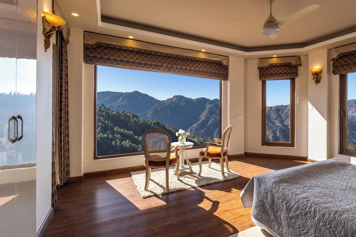 A bedroom with a large window offering a mountain view, a bed, a chair, and a small table with a vase of flowers.