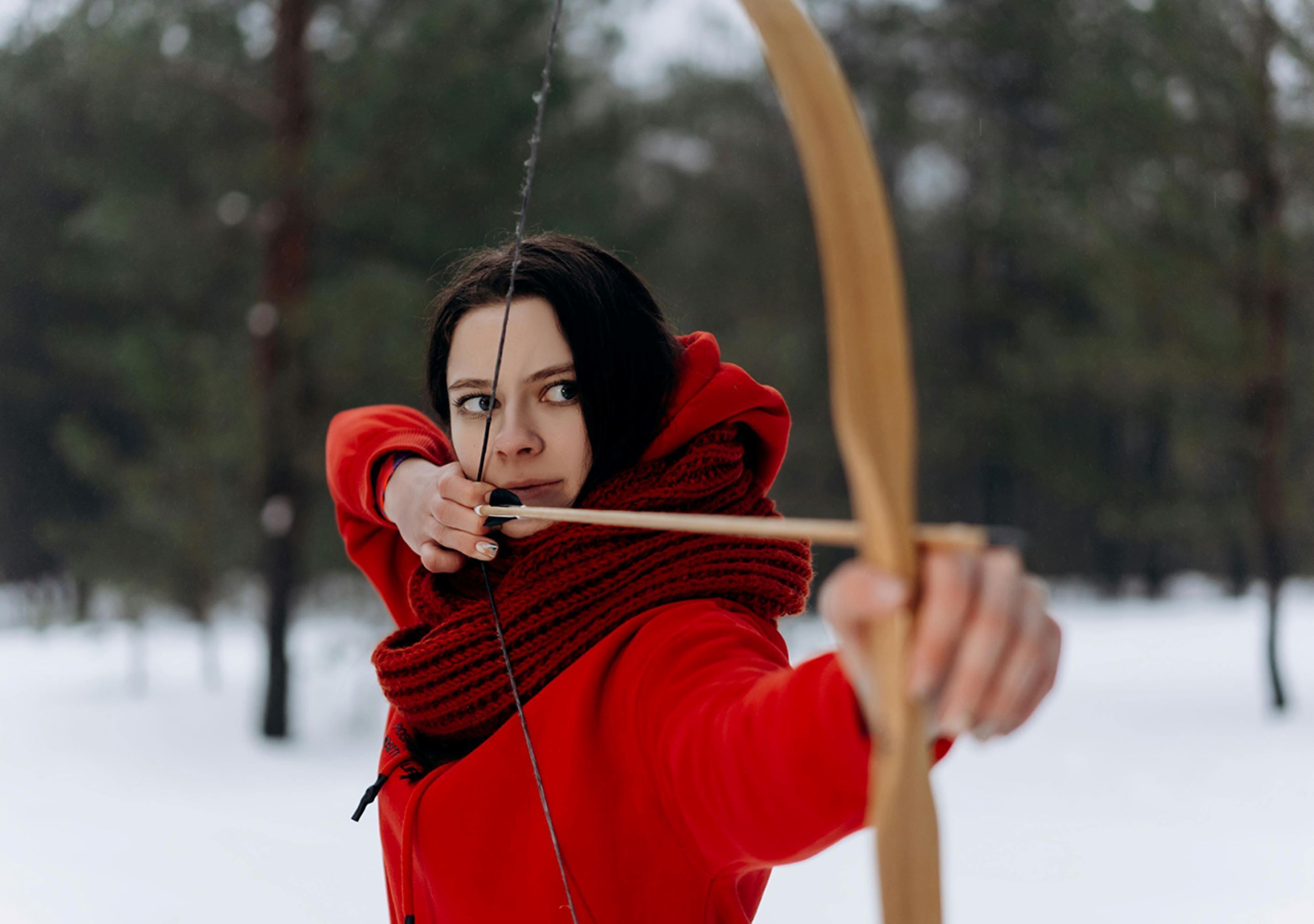 A person in a red jacket aiming a bow in a snowy forest.