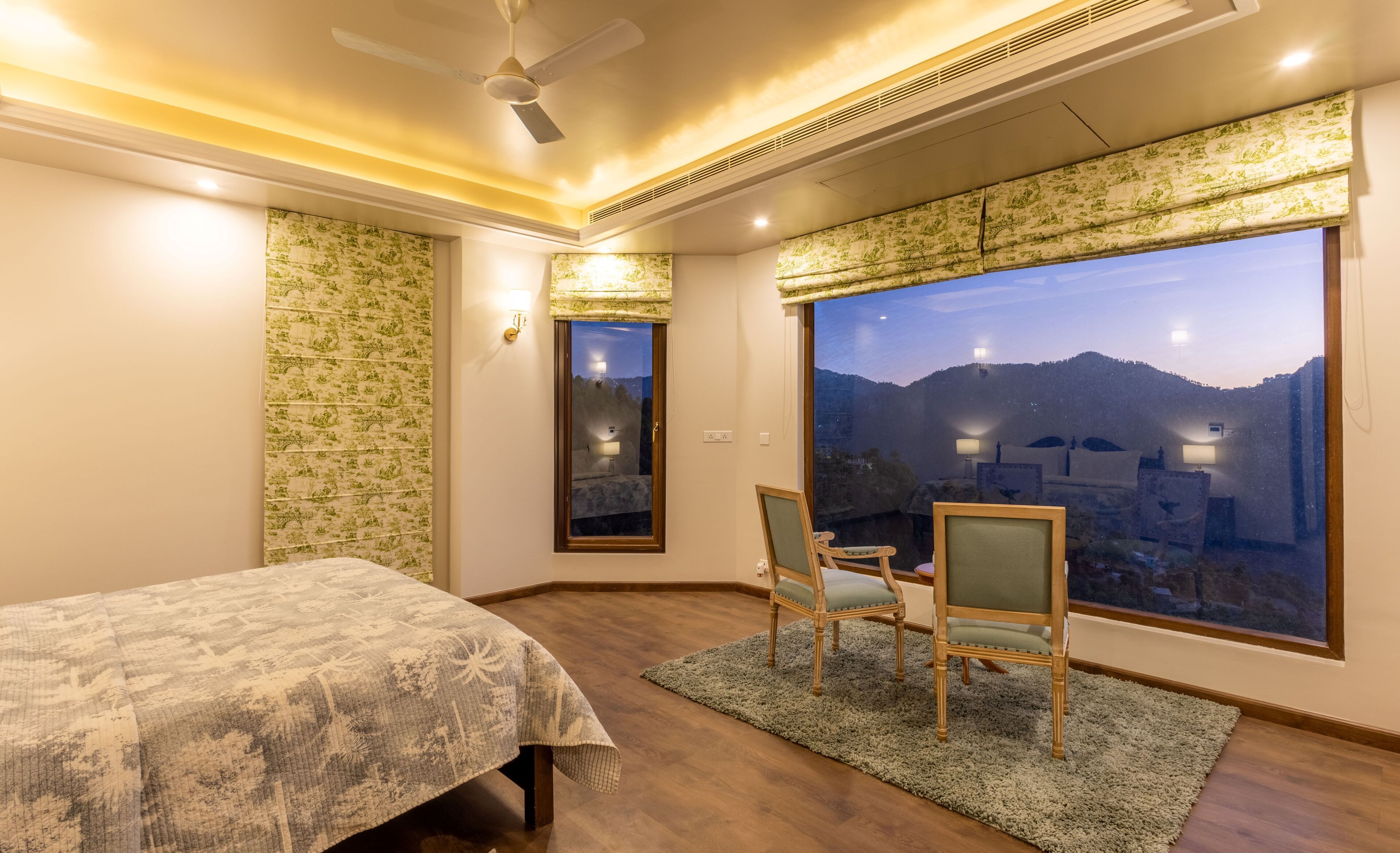 A bedroom with a bed, two chairs, a large window with a view of mountains, and a ceiling fan.