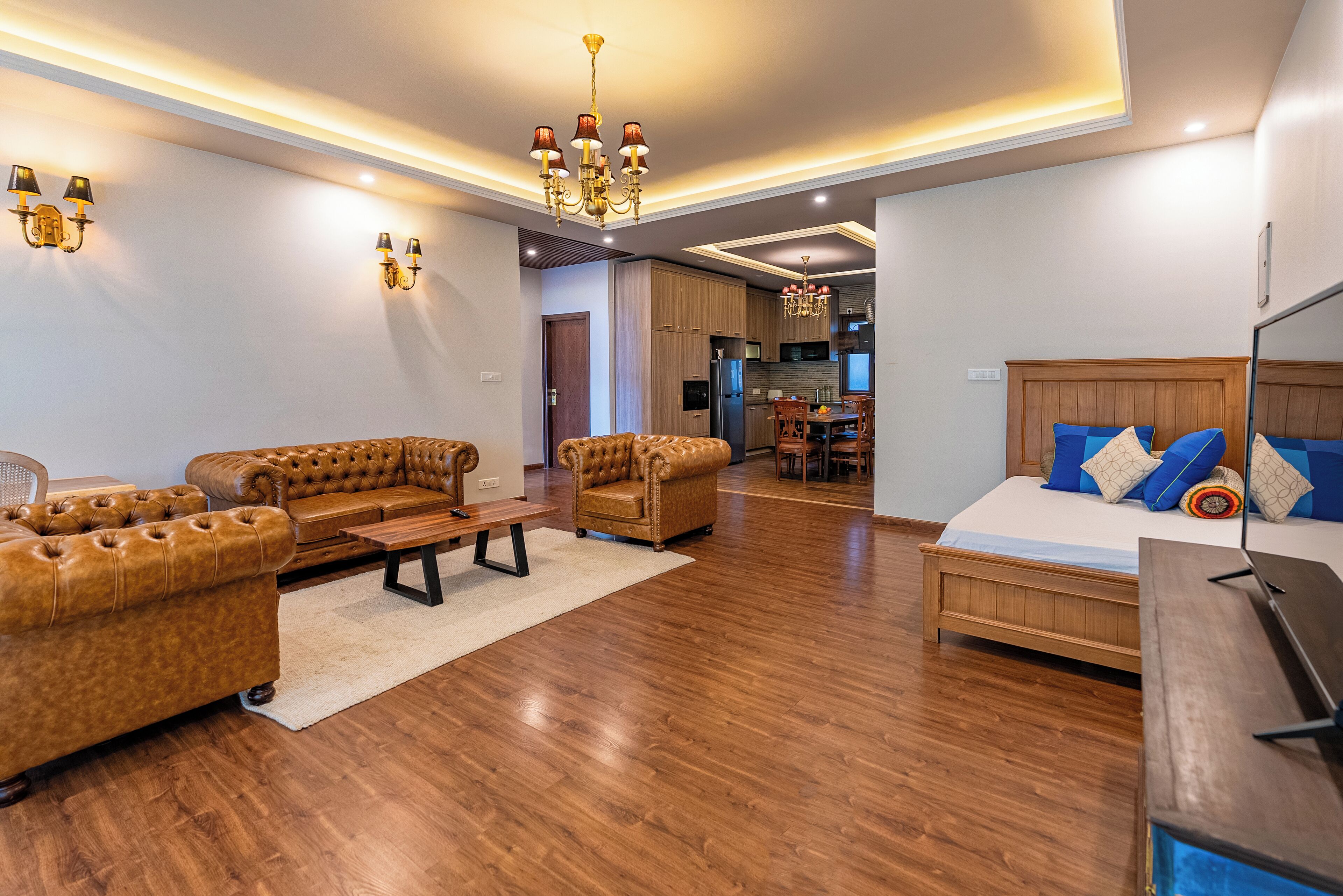 A spacious living area with a sofa, armchair, and a wooden coffee table. There is a bed with blue and beige pillows, and a TV on a wooden stand. The room has wooden flooring and a chandelier.