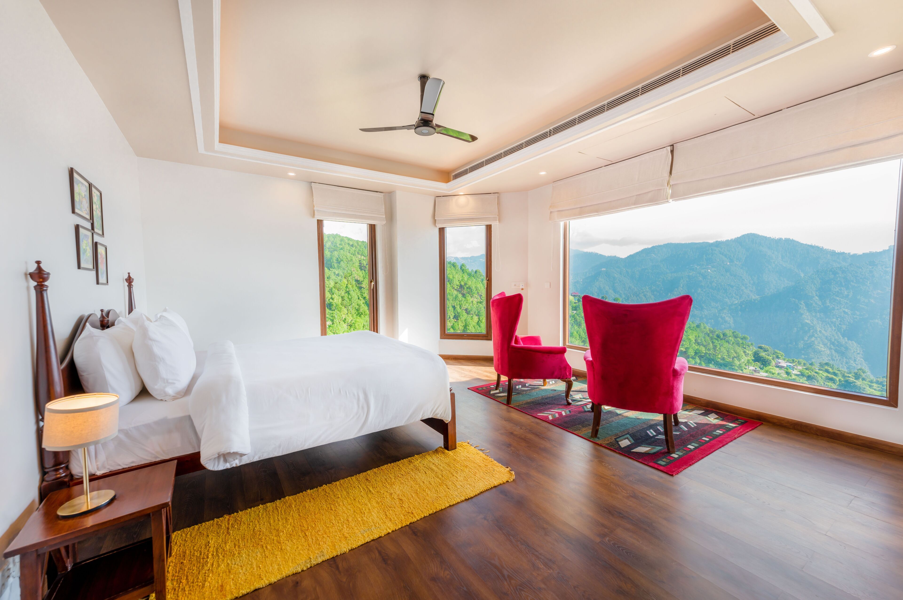 Une chambre à coucher avec un lit, un fauteuil rouge, un tapis jaune et de grandes fenêtres donnant sur la montagne.