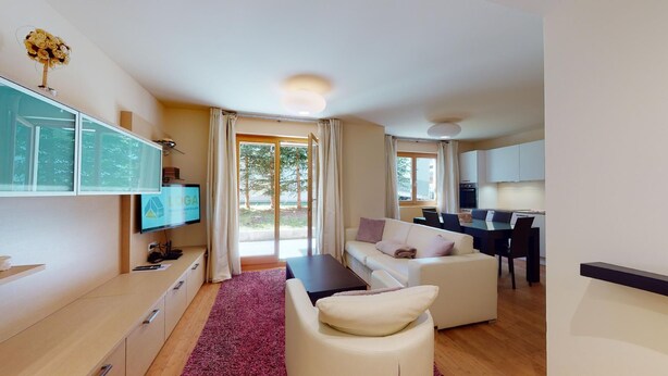 A modern living room with a sofa, a coffee table, and a TV on a wall-mounted stand. There is a dining area with a table and chairs, and a kitchen area with cabinets and a countertop. - Modern 3½-room apartment on the ground floor, approx. (Madulain)