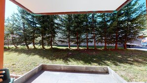 An outdoor patio area with a concrete seating space, surrounded by tall evergreen trees and a view of a body of water. - Modern 3½-room apartment on the ground floor, approx. (Madulain)