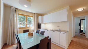 A modern kitchen with white cabinets, a stainless steel sink, and a dining area with a glass table and chairs. - Modern 3½-room apartment on the ground floor, approx. (Madulain)