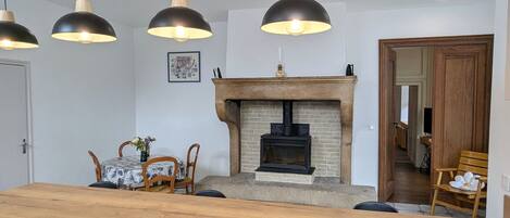 Un espace repas comprenant une table en bois, trois lampes suspendues, une cheminée et une porte donnant sur une autre pièce.