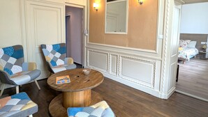 Een kamer met een houten vloer, een ronde houten tafel, twee fauteuils met een gestreept kussen en een bed met een bloemenprint. Er hangt een spiegel aan de muur.