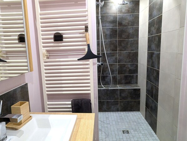 A modern bathroom with a large mirror, a wooden vanity, a shower with a handheld showerhead, and dark tiled walls.