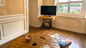 Een kamer met een houten vloer, een televisie op een houten bureau, een naaimachine en een raam met uitzicht op groen.