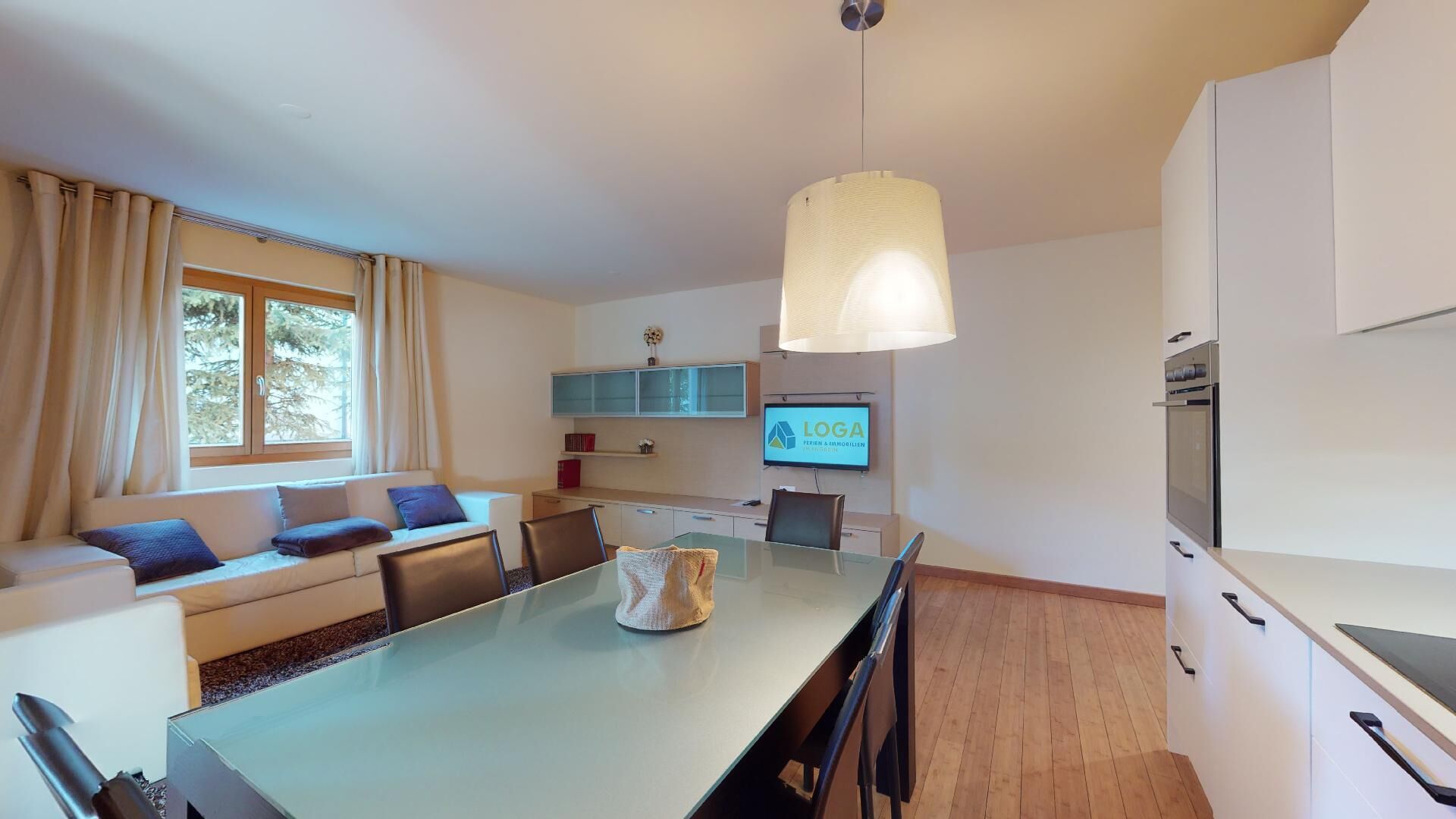 A modern kitchen with a dining table, chairs, a TV, and a large window with curtains.